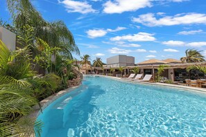 A swimming pool with lounge chairs and palm trees, surrounded by buildings and a clear sky. - NÔA curacao (Willemstad)