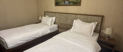 A hotel room with two single beds, a painting of a landscape, a patterned carpet, and a nightstand with a lamp.