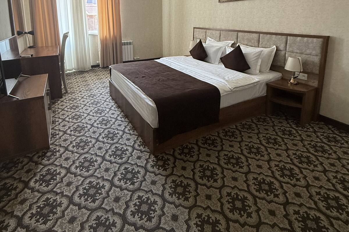 A hotel room with a large bed, a desk, a chair, an air conditioning unit, and a painting on the wall.