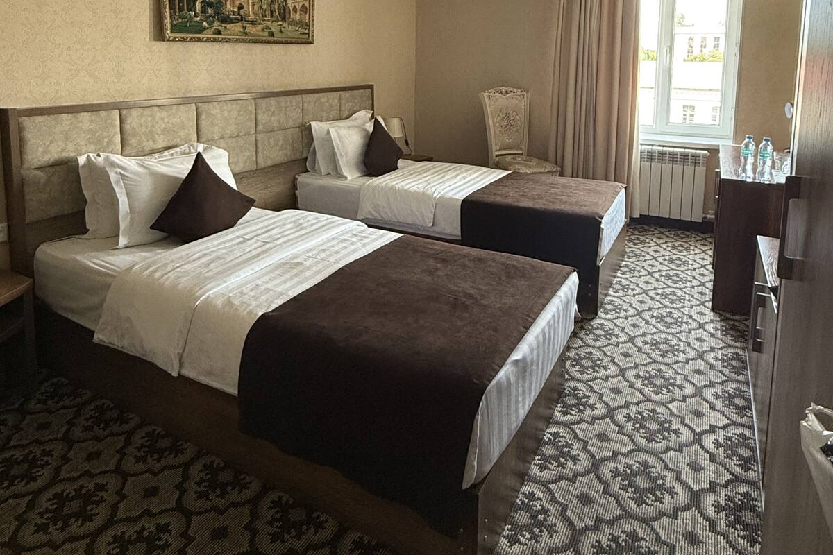 A hotel room with two beds, a chandelier, a painting on the wall, and a window with curtains.