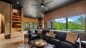 Un salon moderne avec un canapé en cuir noir, une table basse surmontée d’un plateau en fibres tressées et une grande fenêtre donnant sur un paysage verdoyant.