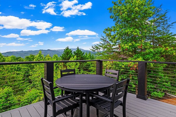 A round table with four chairs on a wooden deck, surrounded by a metal railing, overlooking a lush green landscape with trees and distant hills.