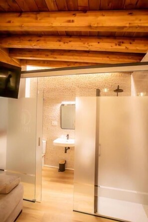 Una habitación con techo de madera, puerta de vidrio que da a un baño con lavamanos y espejo, y un banco acolchado.
