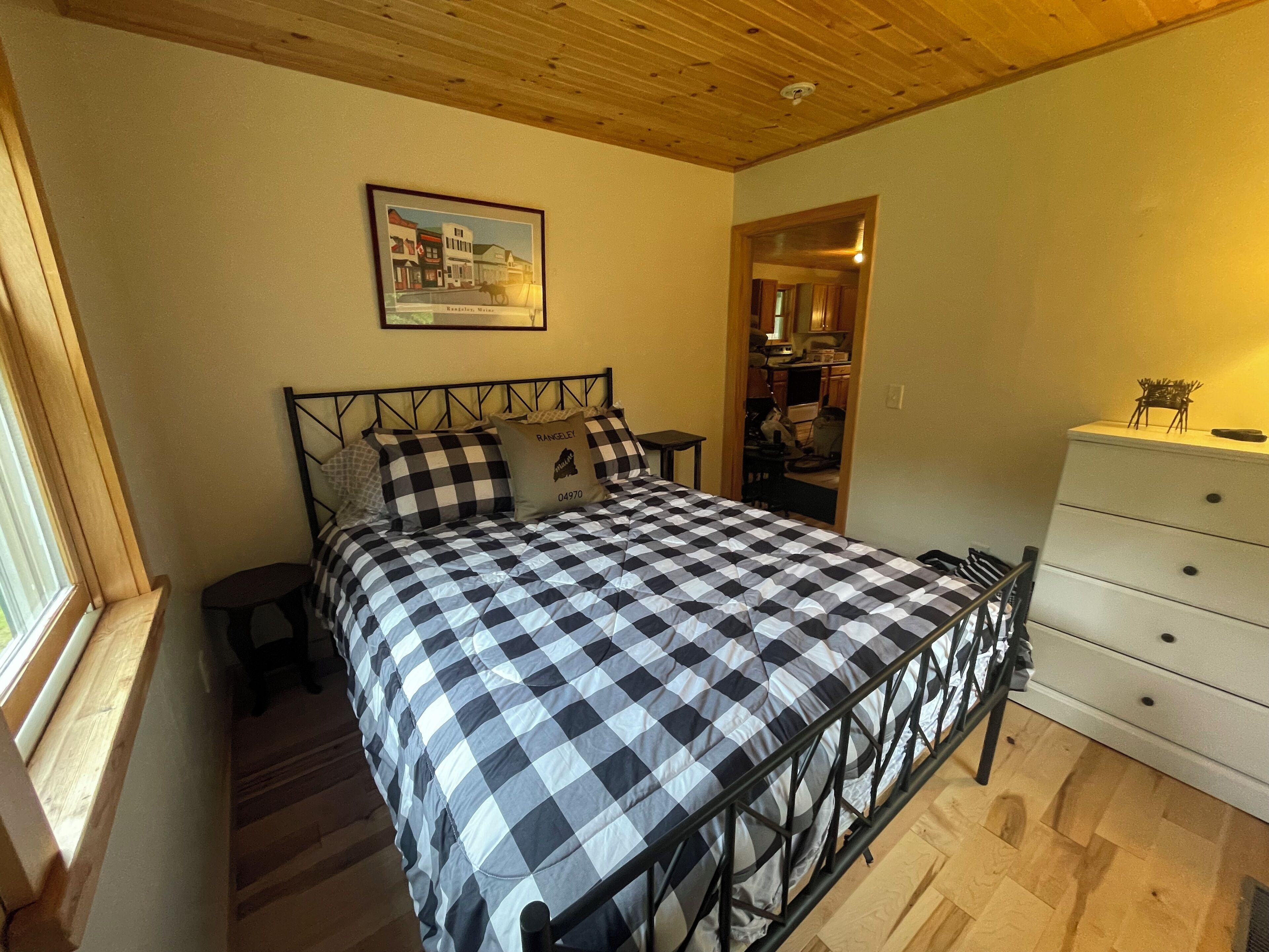 A bedroom with a checkered bedspread, a framed picture on the wall, a dresser, and a window with blinds.