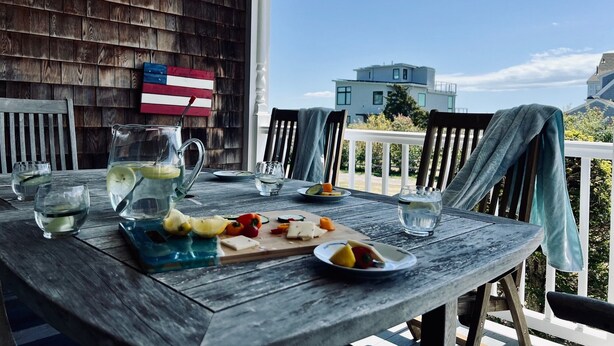 Una mesa de madera con una jarra, vasos y un plato de comida en un balcón con vistas a edificios.