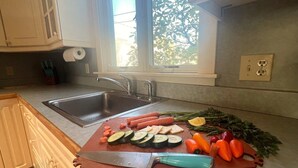Una cocina con un lavamanos, una tabla de cortar con verduras y una ventana con vegetación afuera.