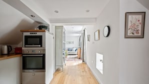 A modern kitchen with a microwave, oven, and kettle. A hallway leads to a living area with a sofa and a rug.
