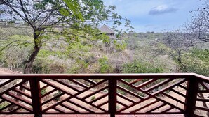 Une terrasse en bois dotée d’une rambarde en treillis, qui offre une vue sur un paysage naturel avec des arbres et un bâtiment situé au loin.