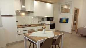A modern kitchen with white cabinets, a stove, and a microwave. There is a dining table with chairs and a vase of flowers. A TV is mounted on the wall. - A&F House - Ale (Torricella)
