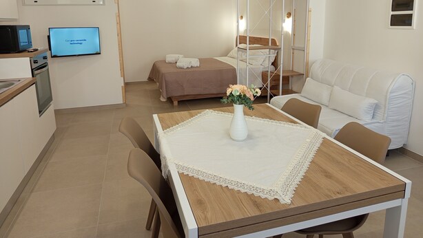 A hotel room with a wooden dining table, a white sofa, a bed with a brown headboard, a TV on the wall, and a small kitchenette. - A&F House - Ale (Torricella)