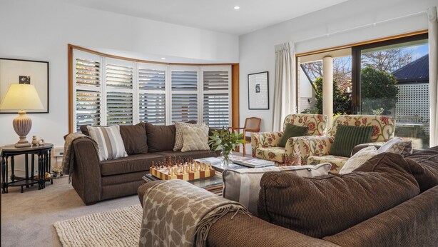A living room with a brown sofa, a floral armchair, a chessboard, and a large window with blinds.