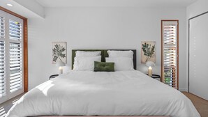 A modern bedroom with a large bed, two bedside tables with lamps, and a view through the window blinds.