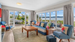 A spacious living area with a large sofa, armchairs, and a coffee table. Large windows offer a view of the sea and greenery. - Herons Hideaway - Snells Beach Holiday Home (Snells Beach)