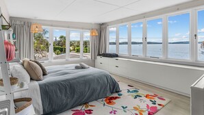 A bedroom with a large window, a bed, a dresser, and a view of a lake. - Herons Hideaway - Snells Beach Holiday Home (Snells Beach)
