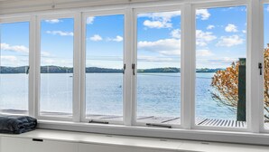 A room with a large window overlooking a lake, a view of distant hills, and a bench with a blanket. - Herons Hideaway - Snells Beach Holiday Home (Snells Beach)