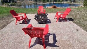 A patio area with red Adirondack chairs, a fire pit, and a wooden table with an umbrella, overlooking a lake and surrounded by trees. - Escape to Nature, Sleep Under The Stars (Zephyr)