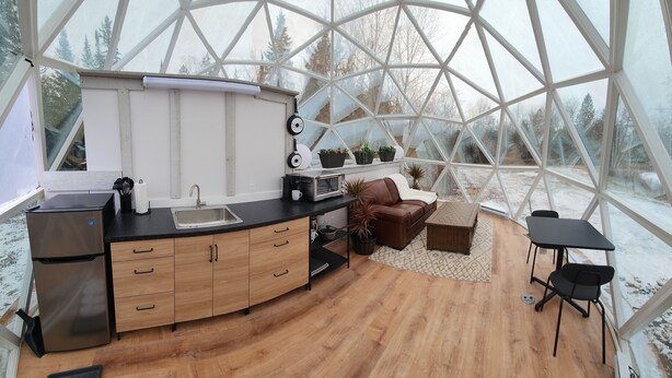 A modern kitchen with a stainless steel refrigerator, wooden cabinets, and a black countertop. There is a sink, a microwave, and a stove. The room has a wooden floor and a dining area with a table and chairs. - Escape to Nature, Sleep Under The Stars (Zephyr)