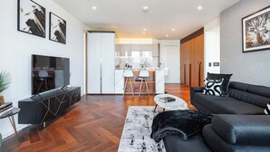 A modern living room with a black sofa, a coffee table, and a TV. There is a kitchen area with white cabinets and a marble countertop. - American Embassy Luxury 2 Bed 2 Bath Top Location! (London)