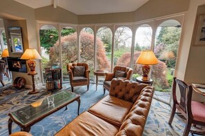 Une pièce chaleureuse avec un canapé en cuir, des fauteuils, une table basse et des lampes. De grandes fenêtres offrent une vue sur la verdure environnante.