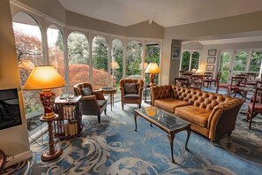 A spacious living room with a large leather sofa, a coffee table, and a fireplace. There are multiple chairs and a dining table in the background. - Burn How Garden Hotel - Bowness on Windermere (Bowness-on-Windermere)