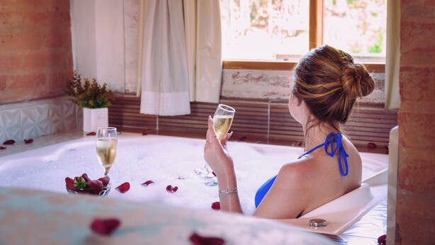 Una mujer en una bañera llena de pétalos de rosa, sosteniendo una copa de champán y un tazón de fresas.