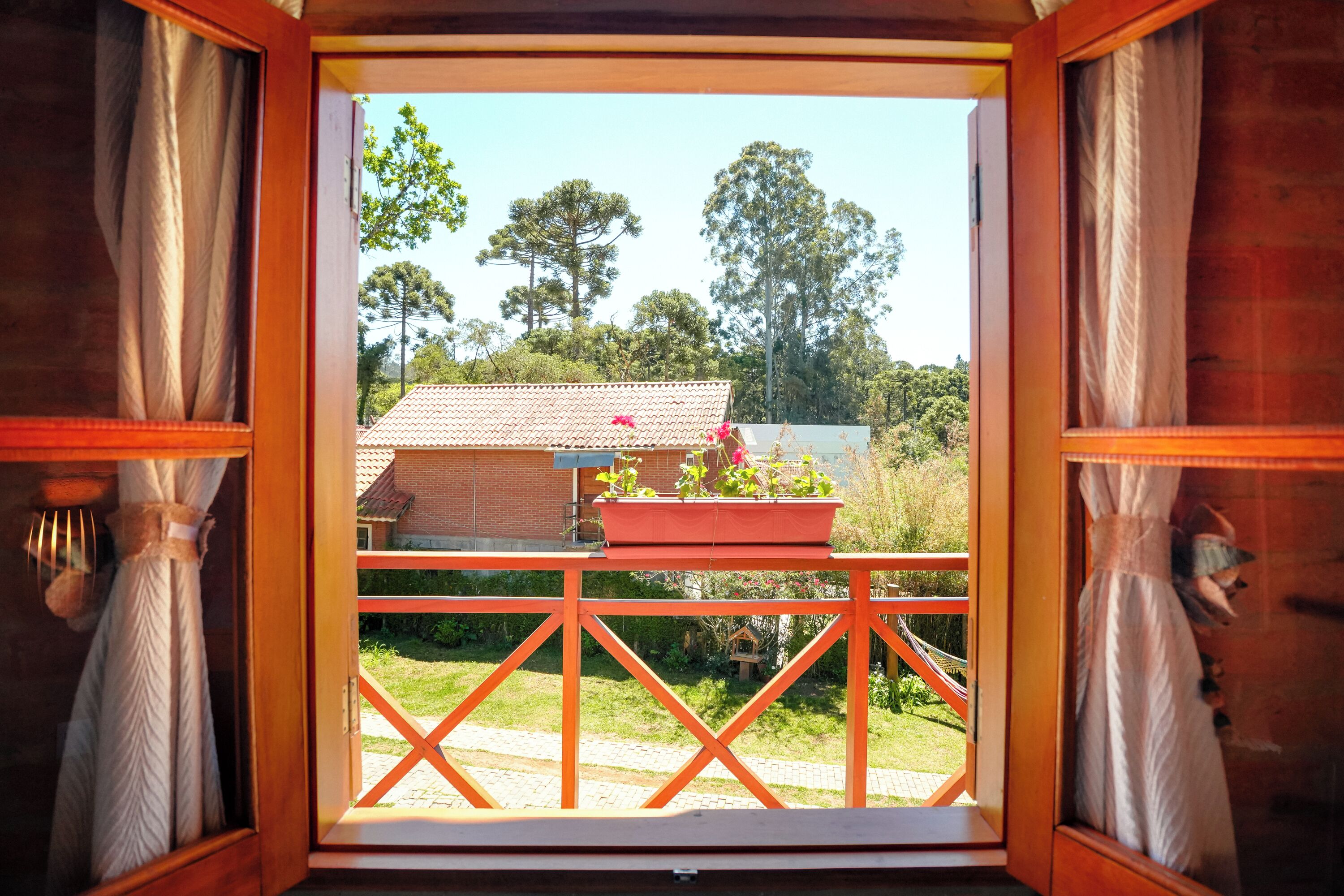 A wooden-framed window with curtains, a balcony with a red railing, and a view of a house and trees.