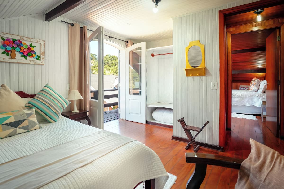Un dormitorio con una cama, un banco de madera, una mesita con una lámpara, una puerta que da a un balcón y un espejo montado en la pared.
