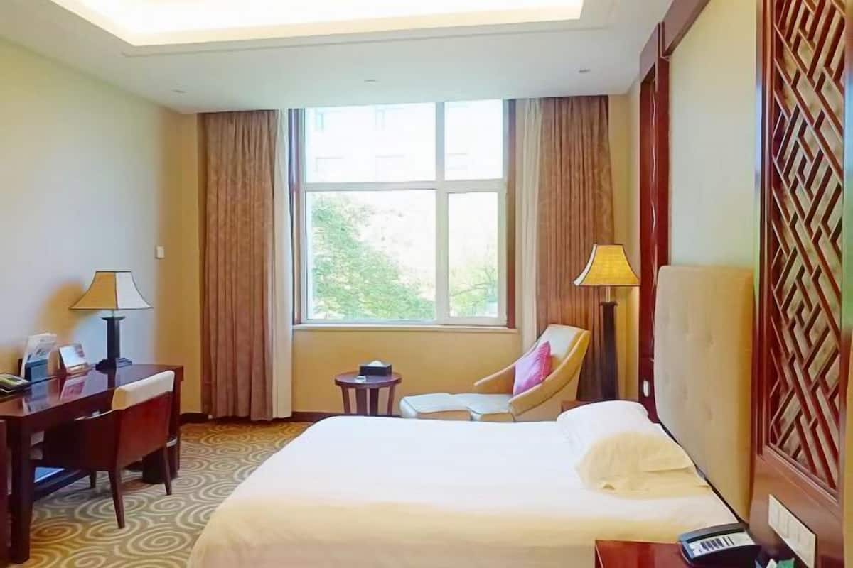A hotel room with a bed, a desk, a chair, a small table, a lamp, and a window with curtains.