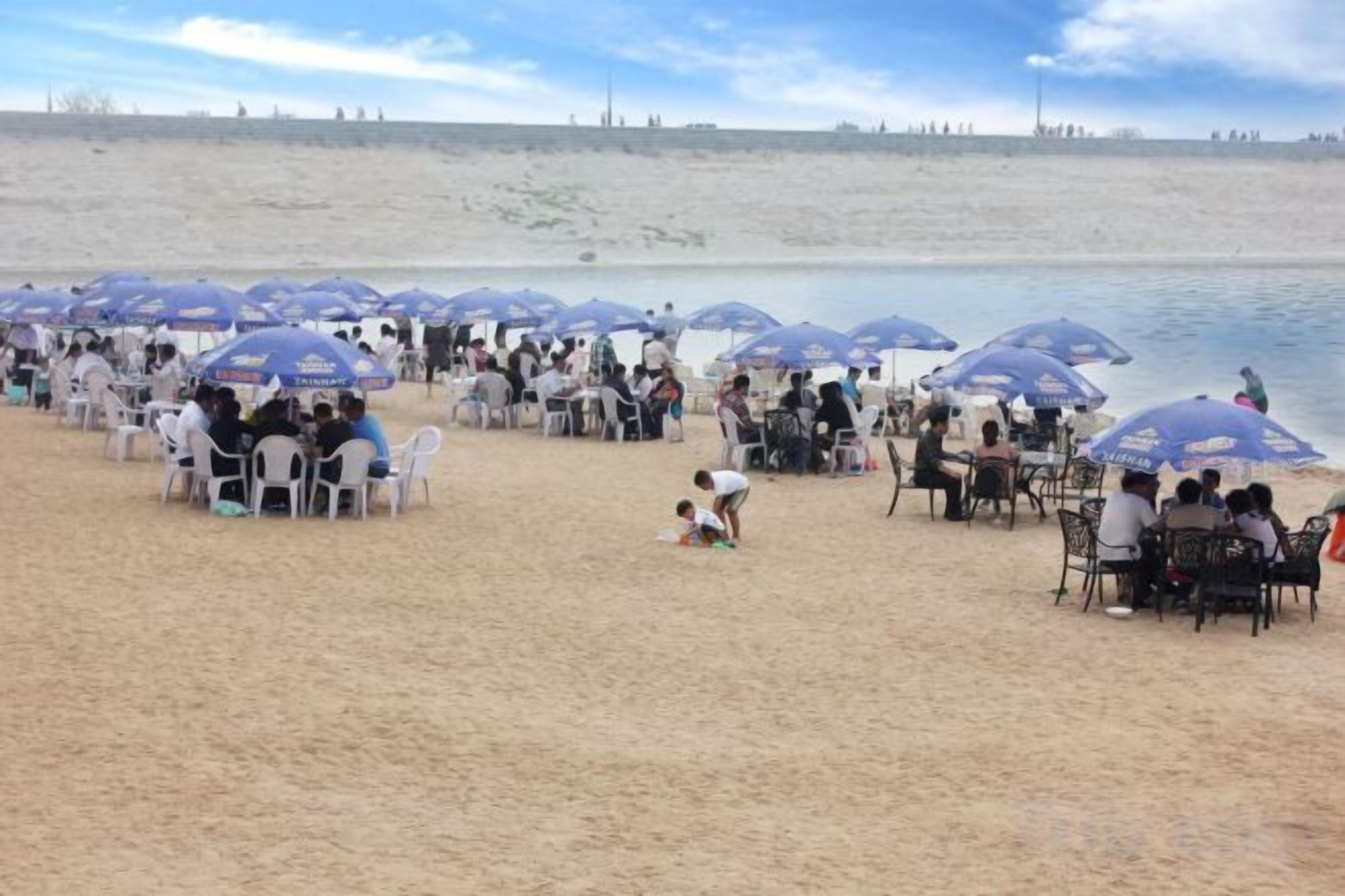 A beach with rows of tables and chairs under umbrellas, people sitting and walking, and a calm body of water in the background.