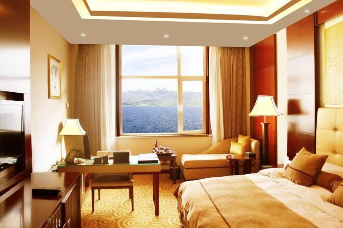 A hotel room with a bed, a desk with a laptop, a chair, a TV, and a view of the sea.