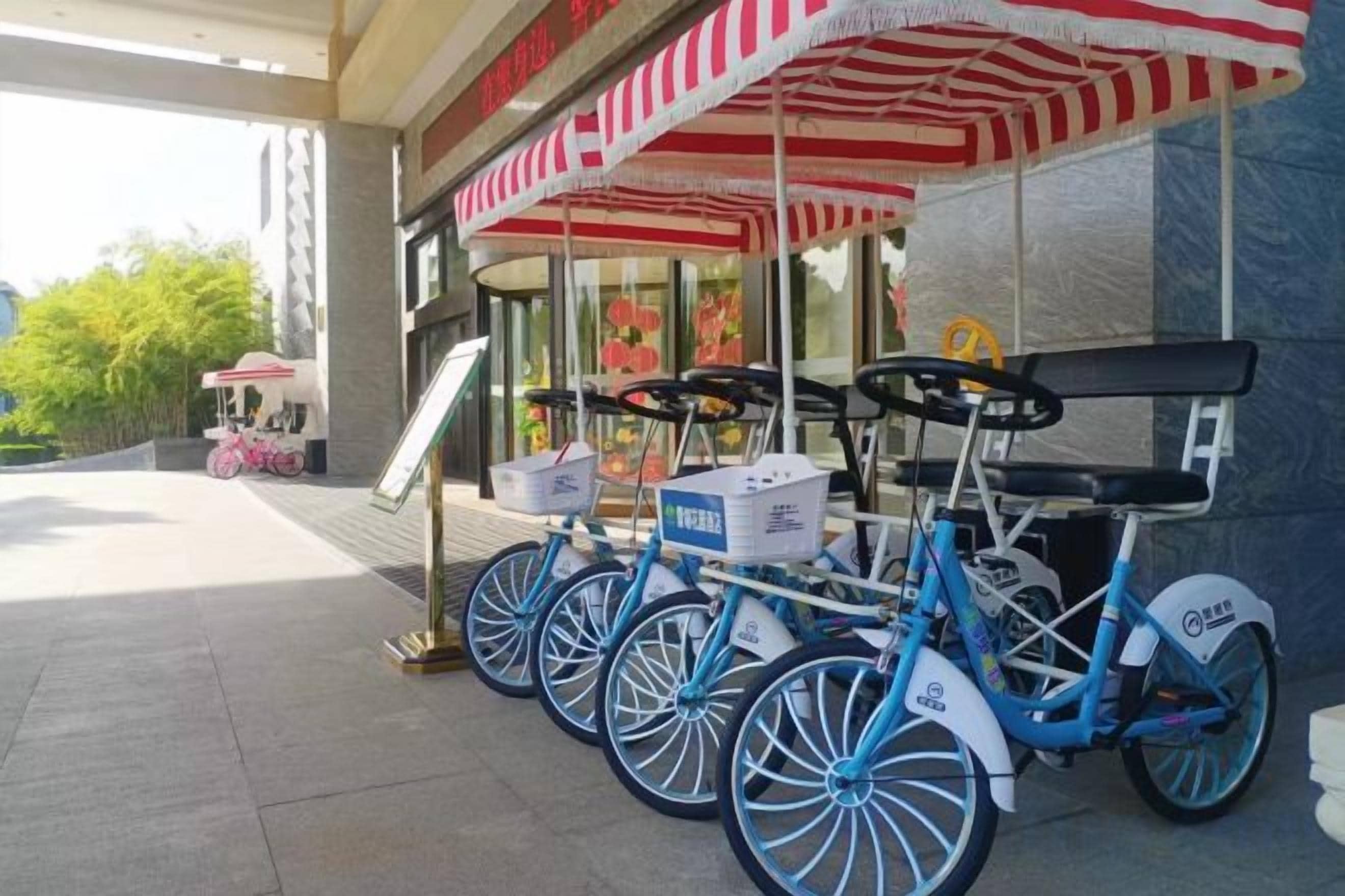 A row of blue bicycles parked under a red and white striped awning, with a signboard and a pink bicycle in the background.