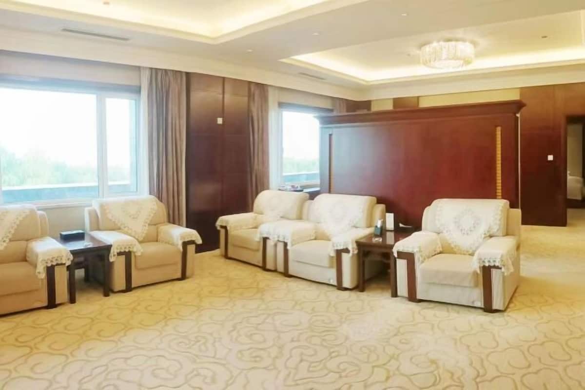 A spacious living room with a patterned carpet, a large window with curtains, and a chandelier.