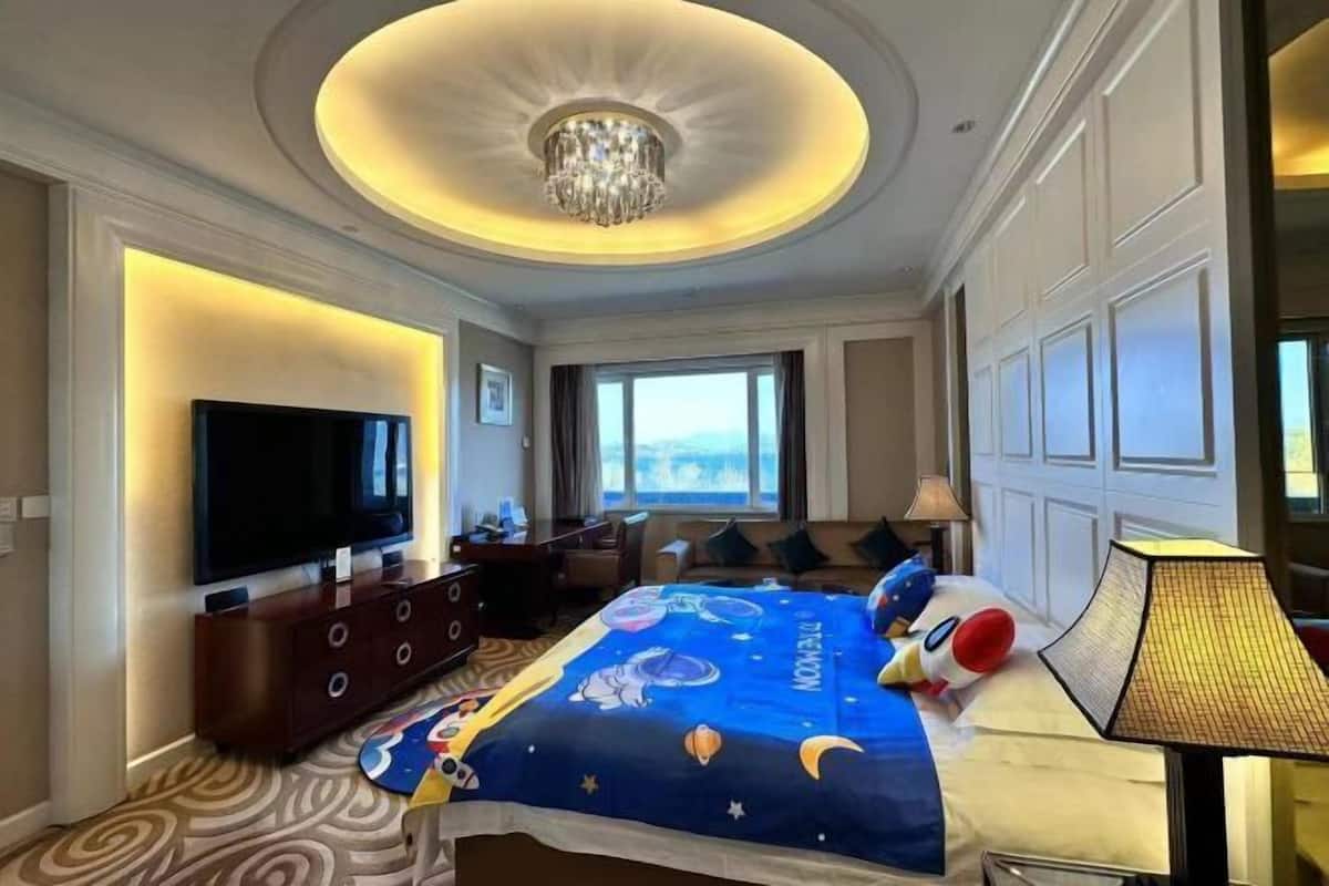 A hotel room with a large flat-screen TV, a bed with a space-themed bedspread, a chandelier, and a view of the outdoors.