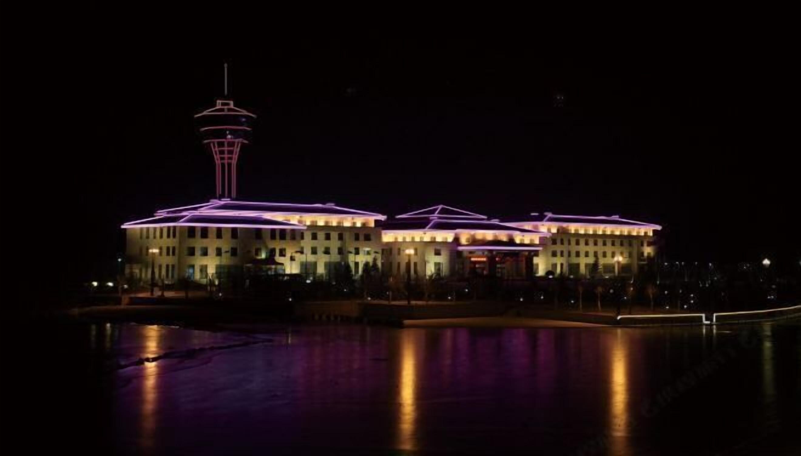 A large building with a tower, illuminated by purple lights, reflecting on a body of water.