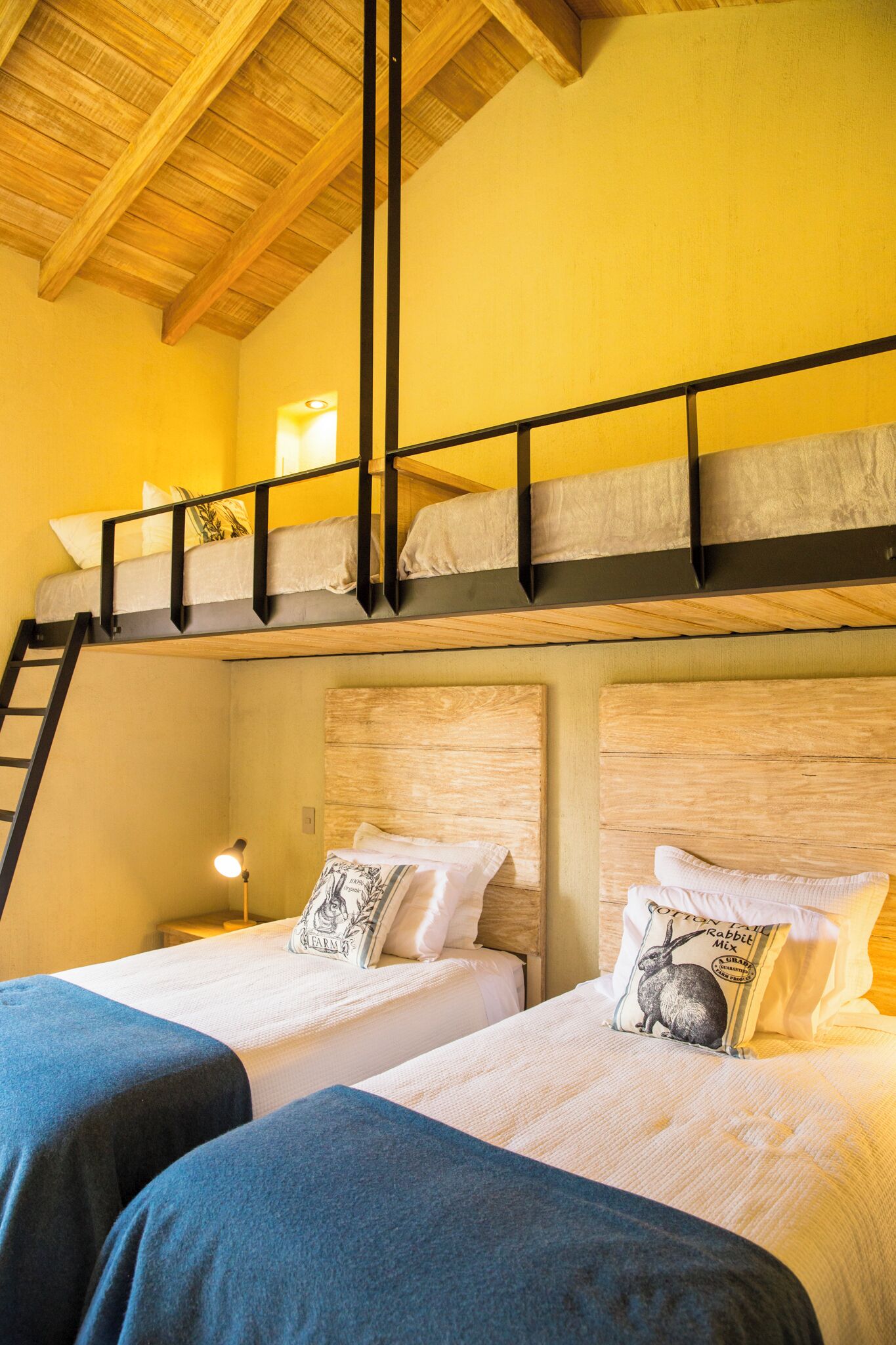 Una stanza con due letti, un letto a castello in alto, travi in legno a soffitto e una scala a sinistra.