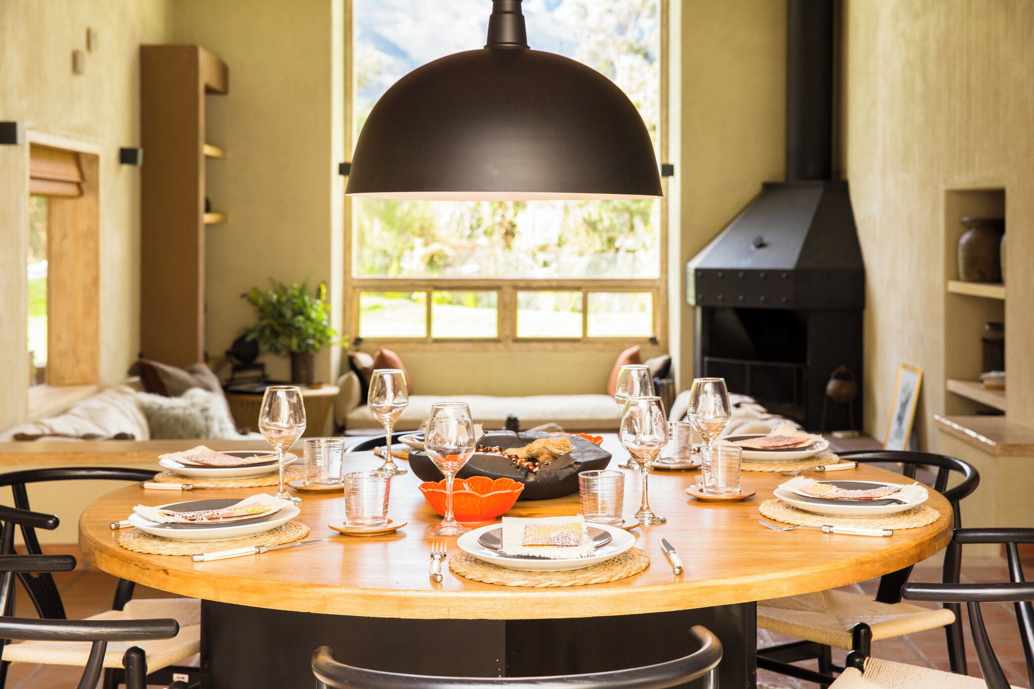 Une table de salle à manger ronde en bois, mise pour un repas, avec des assiettes, des verres et un saladier. Un luminaire noir en forme de dôme est suspendu au-dessus.