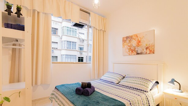 Um quarto bem arrumado com cama, mesa de cabeceira com abajur, janela com cortinas, uma obra de arte na parede e guarda-roupa com roupas penduradas.