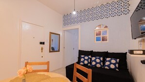 Um quarto com uma mesa de jantar, cadeiras, um sofá preto com almofadas estampadas, um espelho e detalhes decorativos no teto.