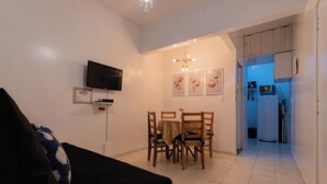 Uma sala de estar com um sofá preto, uma mesa de jantar com cadeiras, uma TV fixada na parede e uma cozinha visível por uma porta aberta.