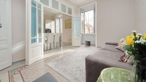 A spacious living room with a sofa, a coffee table, and a vase with flowers. There is a dining area visible through an open door.