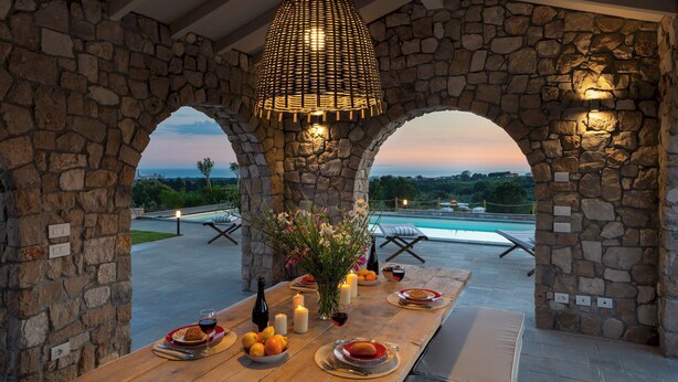A stone-arched outdoor dining area with a pool view, candles, and a bottle of wine.