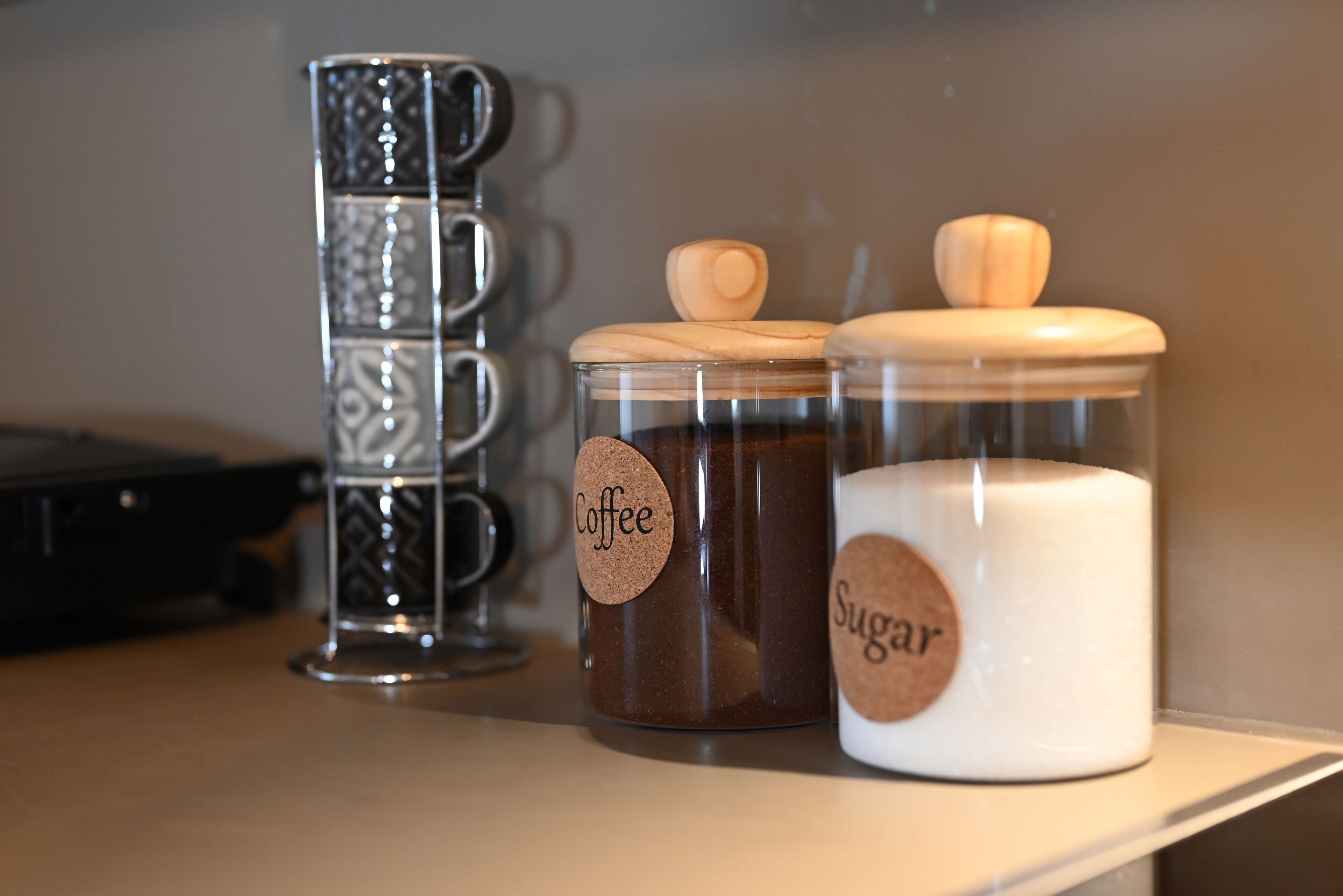 Três recipientes numa prateleira: um para café, um para açúcar e uma caneca preta.