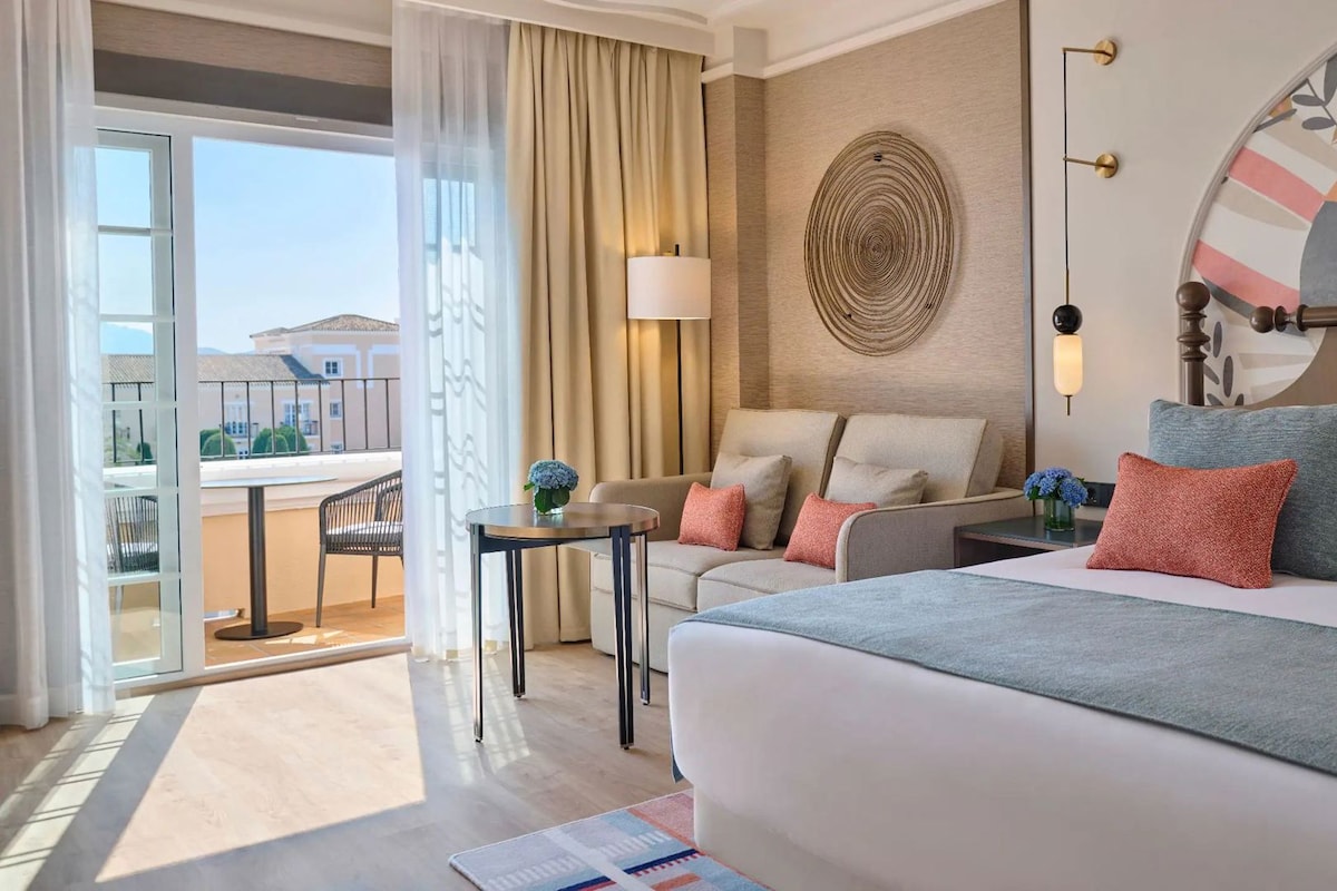 Ein Hotelzimmer mit Bett, Sofa, einem kleinen Tisch, einer Lampe und Blick ins Freie.