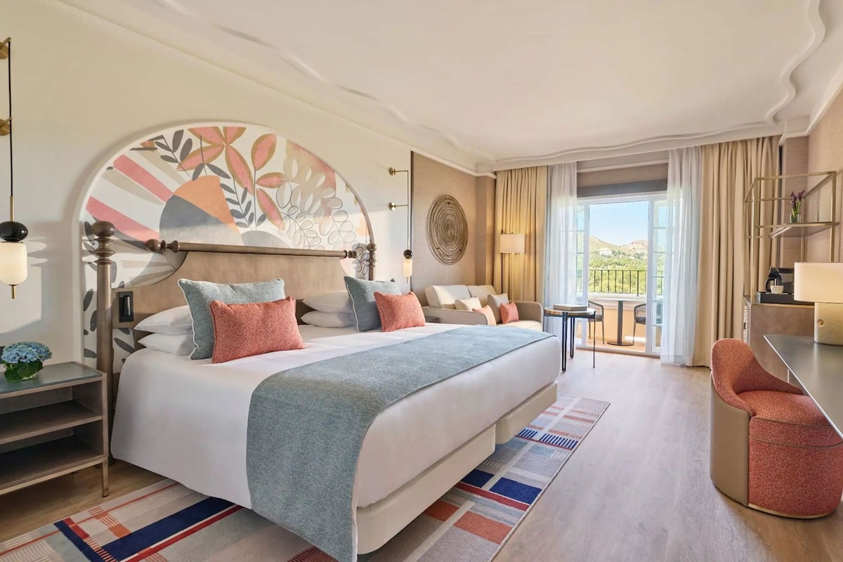 Ein Hotelzimmer mit einem großen Bett, einem Schreibtisch, einem Stuhl und einem Balkon mit Blick.