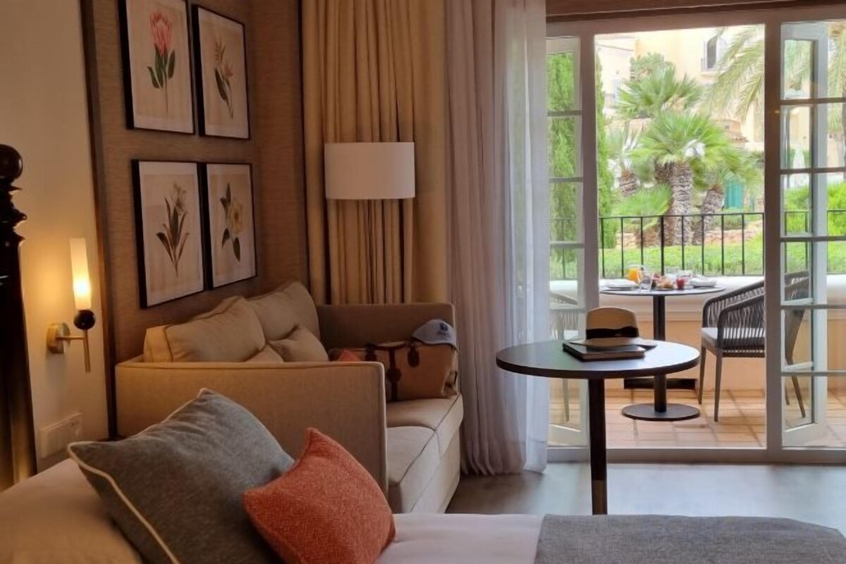 A hotel room with a sofa, a table with a hat, and a view of a garden through a glass door.