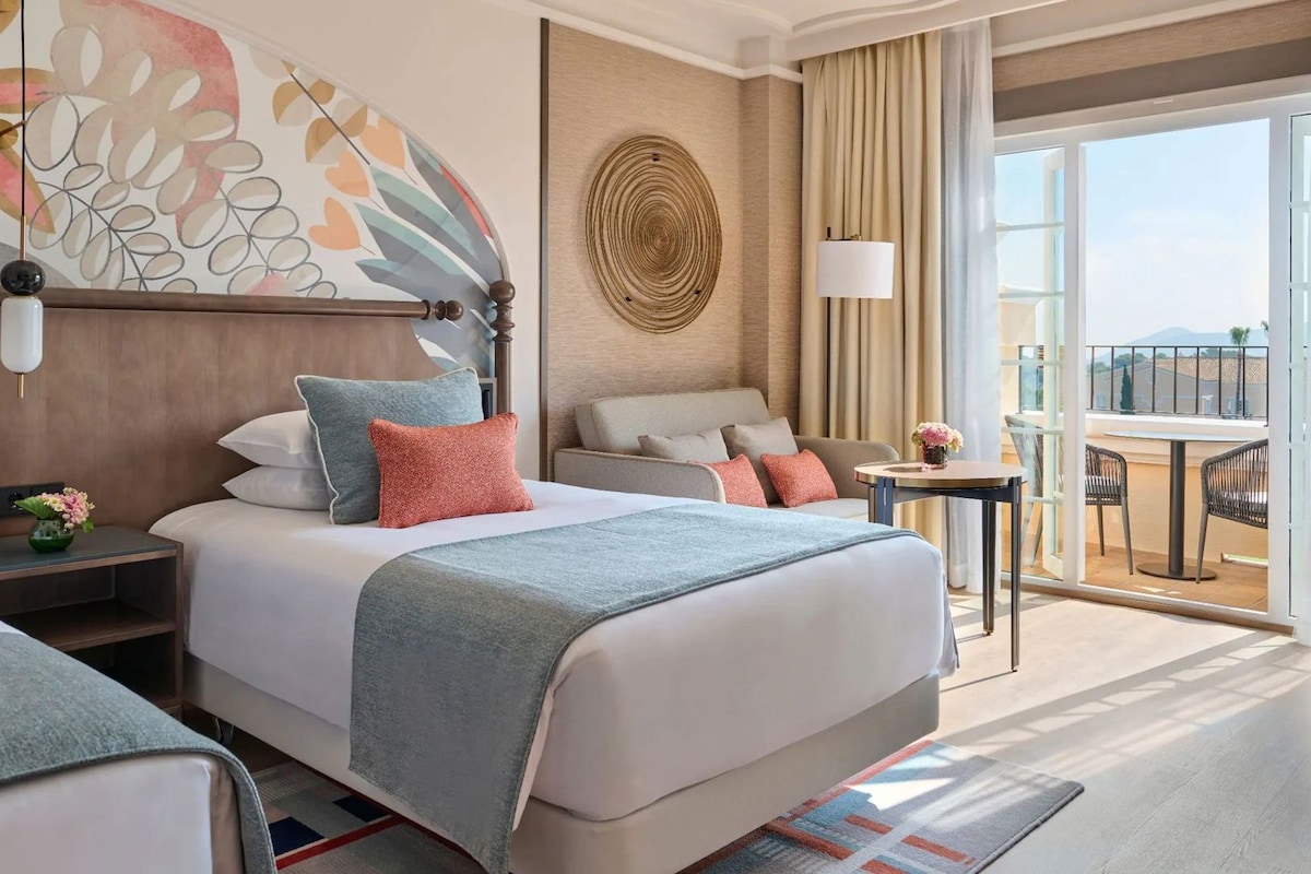 Ein Hotelzimmer mit Bett, Nachttischen, einem Schreibtisch, einem Sessel und einem Balkon mit Tisch und Stühlen, sowie einem großen Fenster.