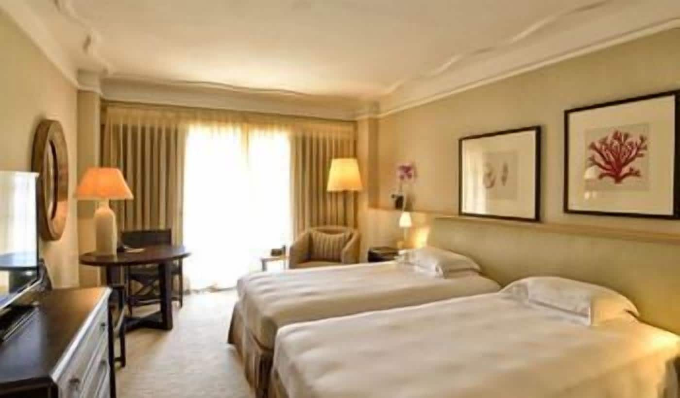 A hotel room with two beds, a desk, a chair, and a lamp.