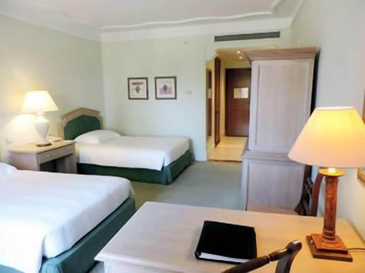A hotel room with two beds, a bedside table with a lamp, a chair, and a door leading to another room.