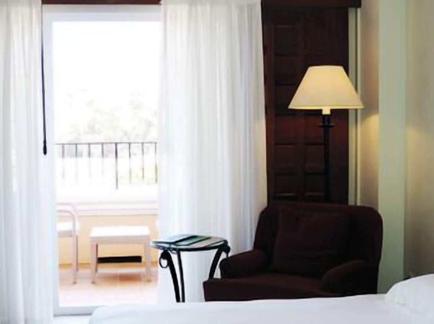 Habitación de hotel con cama, una silla, una mesa y una lámpara. Tiene un balcón con una silla y una mesa.