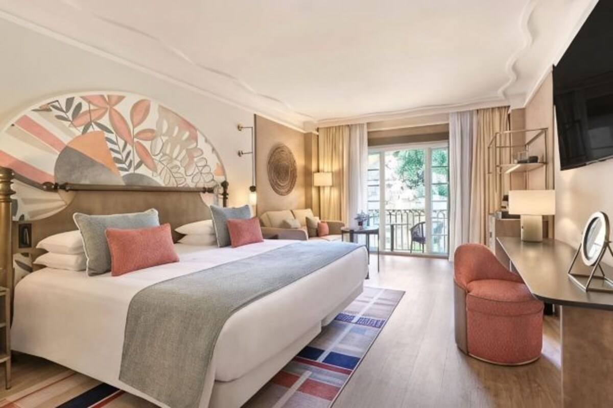 Ein Hotelzimmer mit einem großen Bett, einem Schreibtisch, einem Sessel, einem Fernseher und einem Balkon mit Blick.
